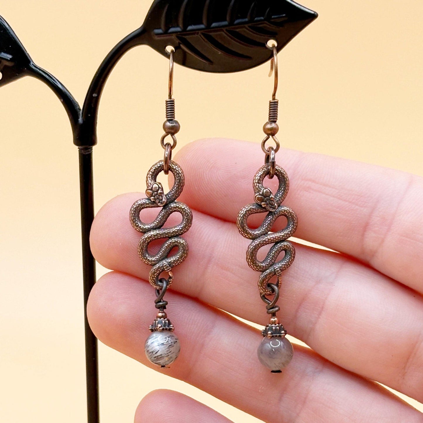 Pair of bronze chain earrings with pearl-like beads held by a hand against a yellow background
