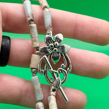 Close-up of a hand holding a beaded bracelet with a silver flower toggle against a green background
