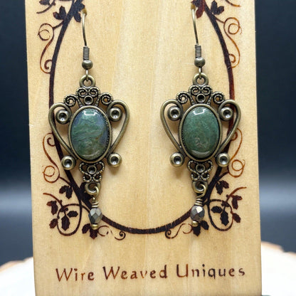 Pair of ornate earrings with green stones on a wooden display card with 'Wire Weaved Uniques' branding.