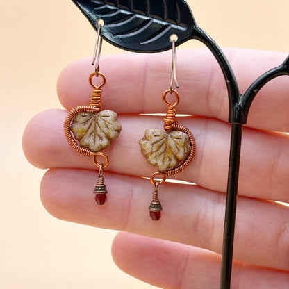Pair of leaf-shaped earrings with wire wrapping held between fingers against a beige background