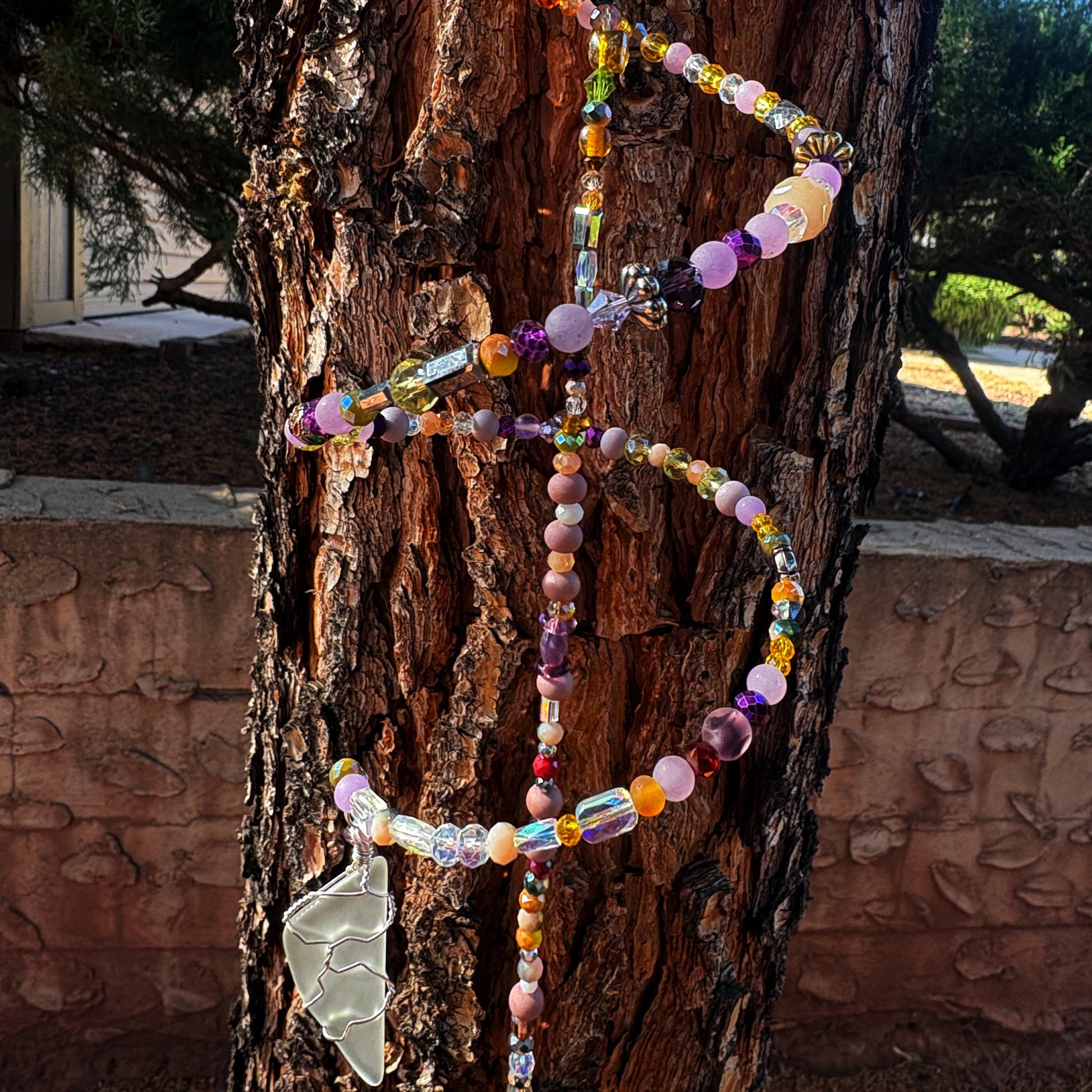 Colorful beaded sun catcher with a wire wrap sea glass pendant hanging on a tree trunk.
