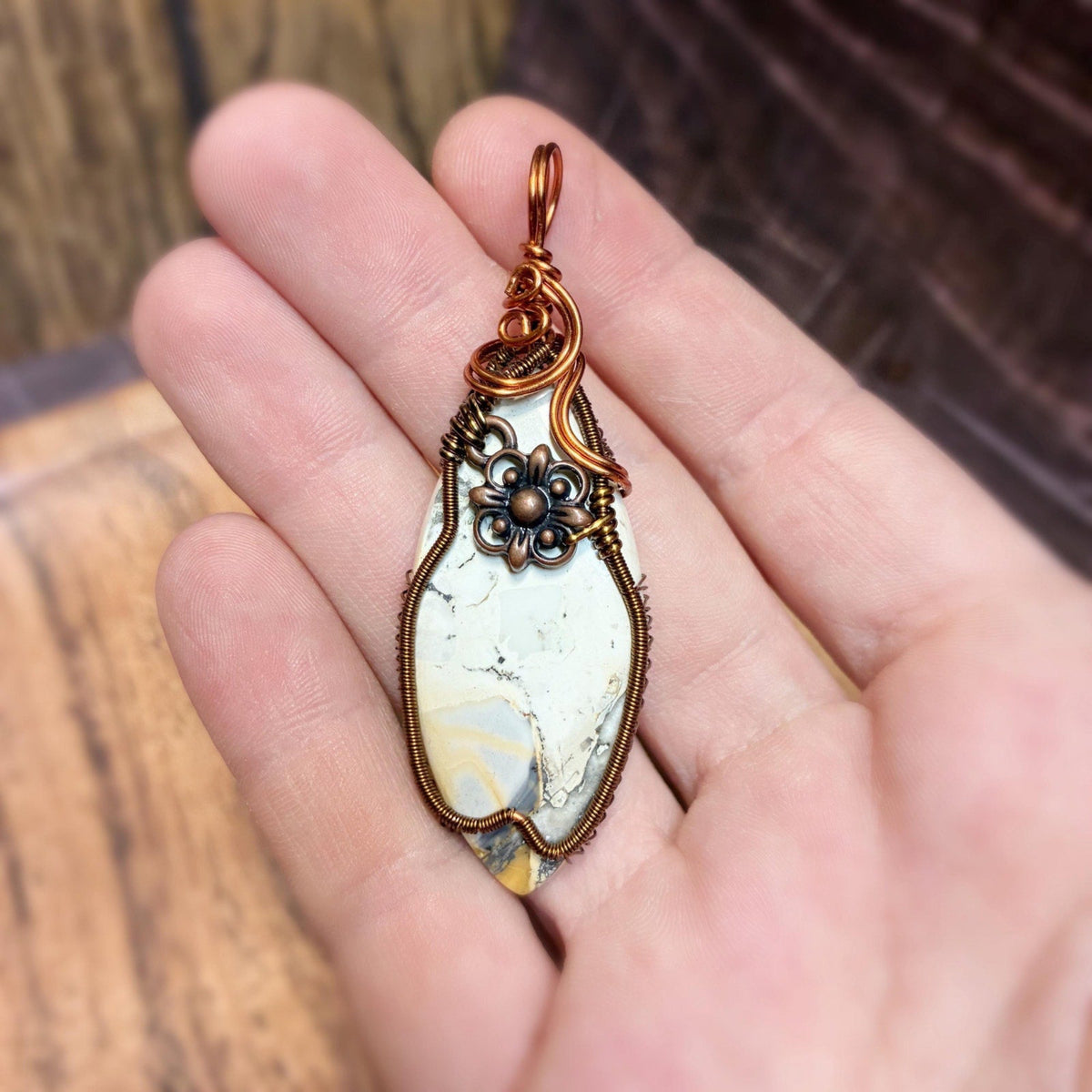 A hand holding a marquise-shaped pendant made from a white marbled stone with copper wire wrapping and a small black floral accent.