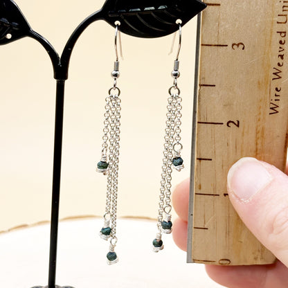 Pair of silver chain earrings with green beads on a black earring stand next to a wooden ruler.