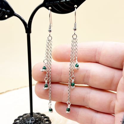 Silver chain earrings with green beads held by a hand against a neutral background