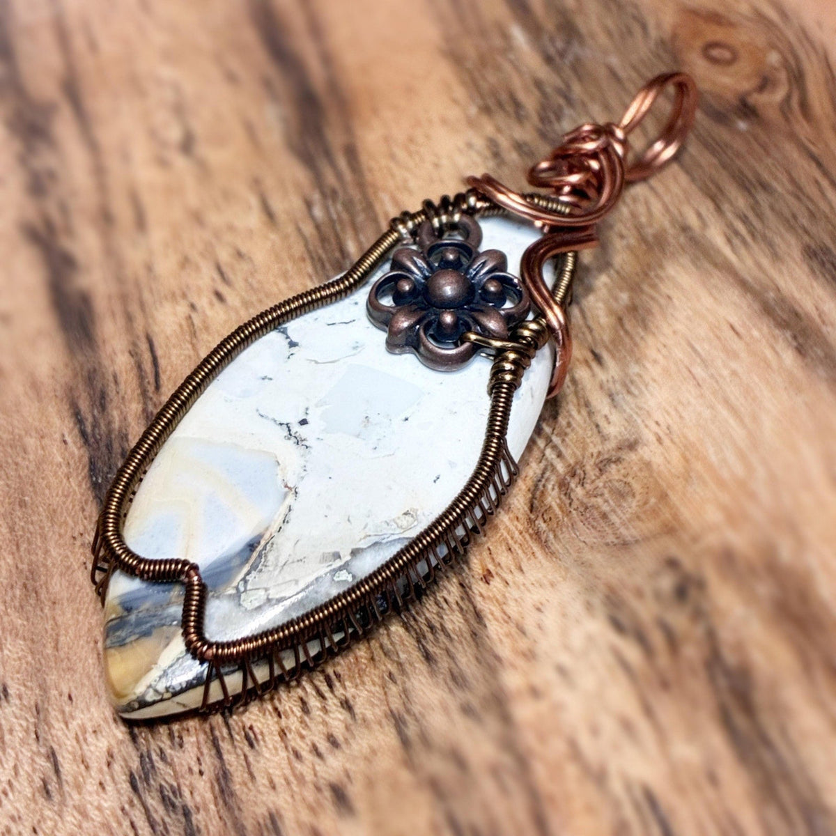 A handmade wire-wrapped pendant featuring a white and gray marbled stone with copper-toned metal wrapping and a dark bronze flower accent, displayed on a rustic wooden surface.