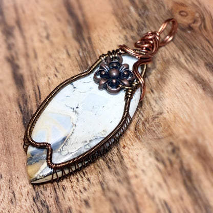 A handmade wire-wrapped pendant featuring a white and gray marbled stone with copper-toned metal wrapping and a dark bronze flower accent, displayed on a rustic wooden surface.