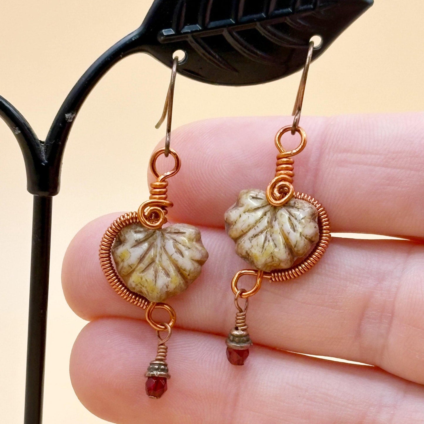 Pair of leaf glass and copper wire earrings held between fingers against a beige background
