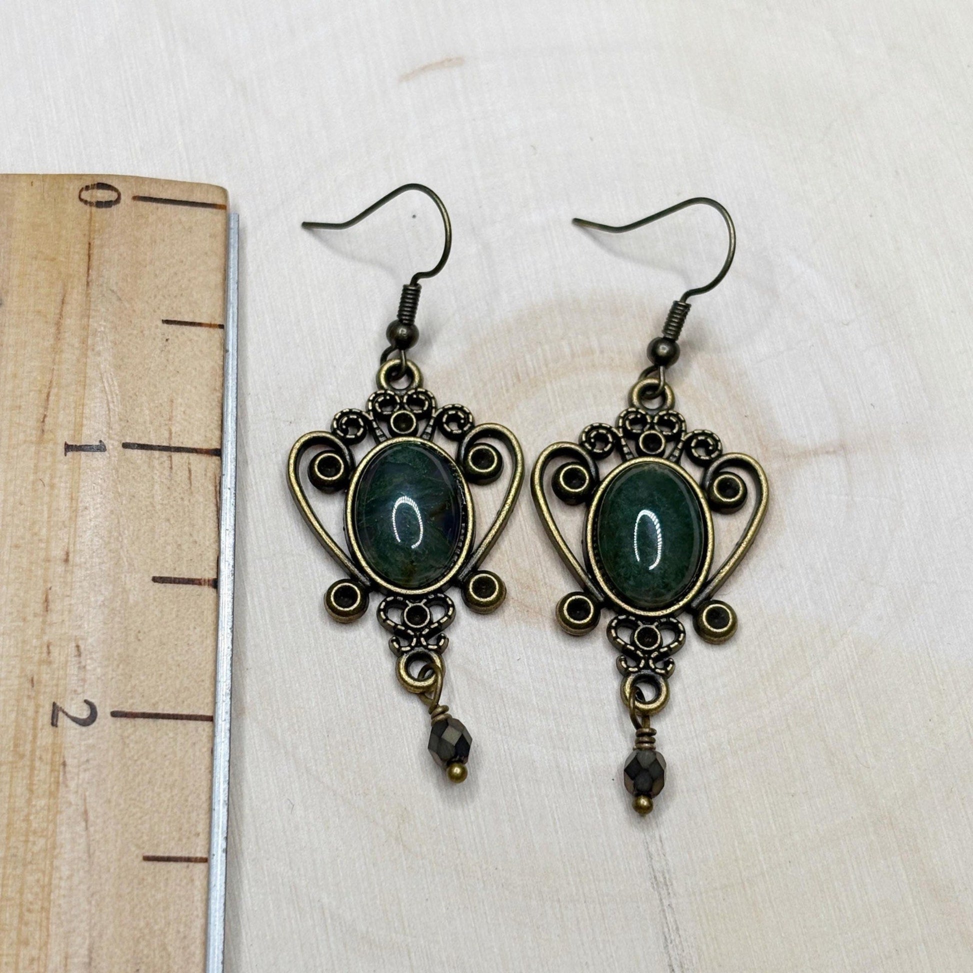Pair of ornate earrings with green stones on a white background with a ruler for scale.