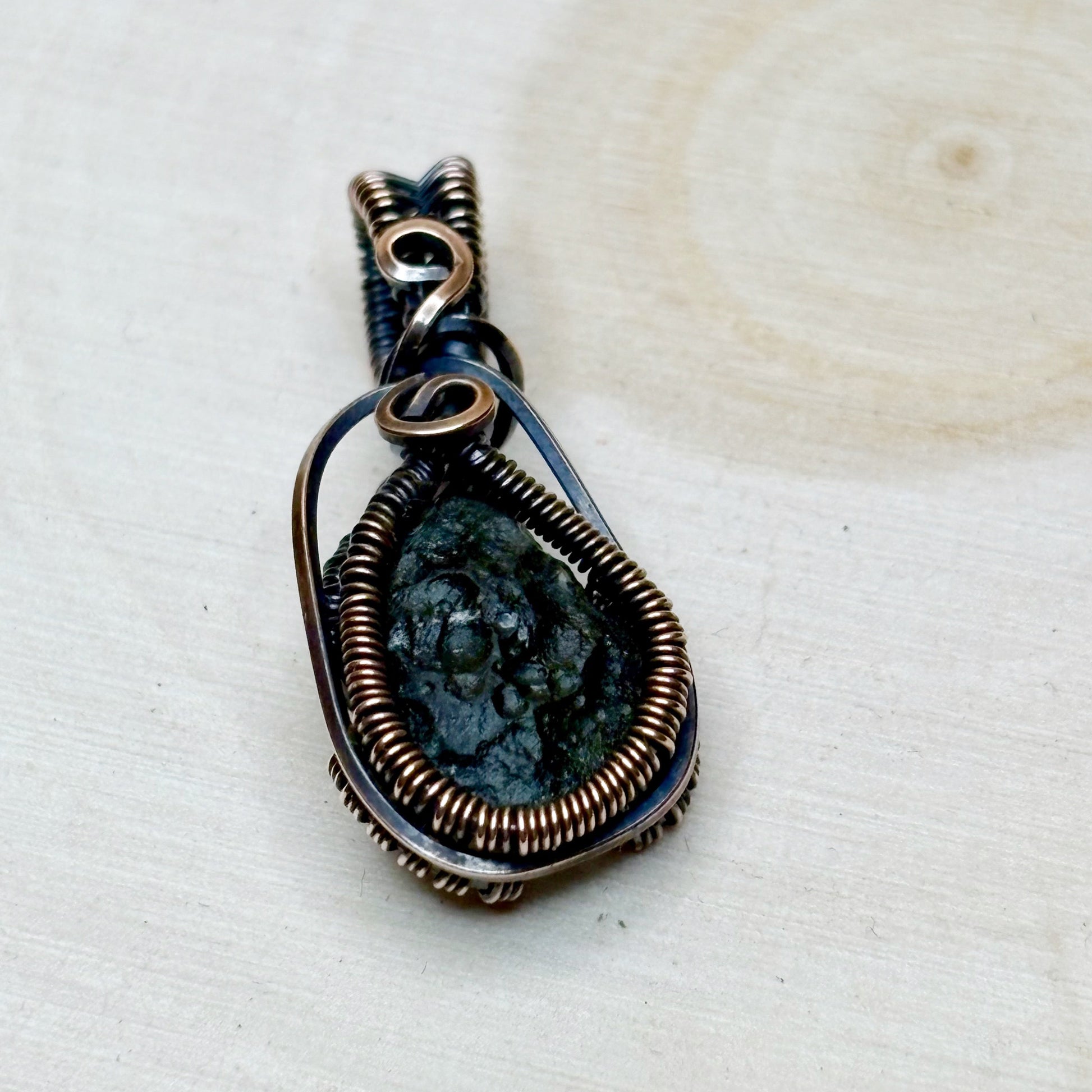 Pendant with a dark green stone and copper wire wrapping on a beige background