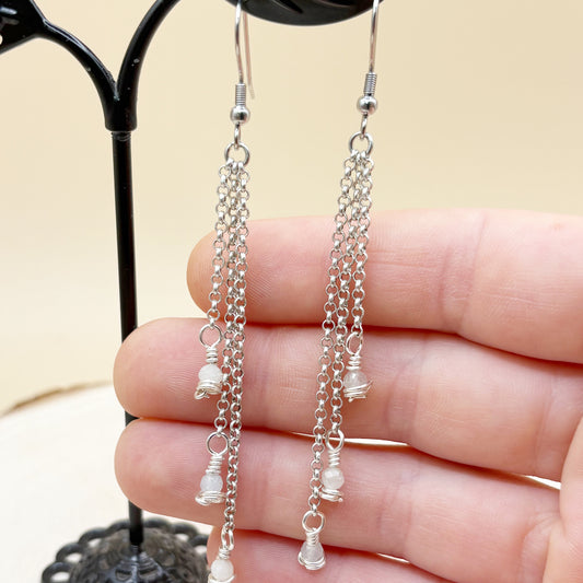 Silver chain earrings with small beads held between fingers against a neutral background