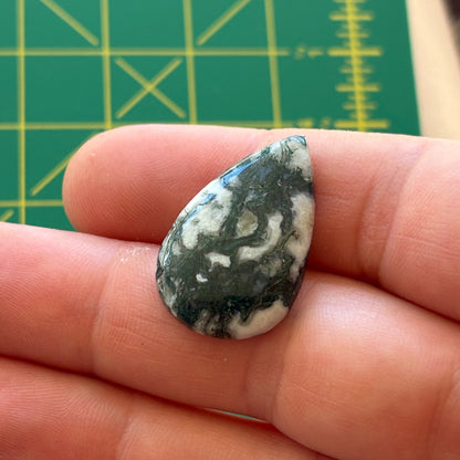 Teardrop-shaped stone held between fingers with a green cutting mat in the background