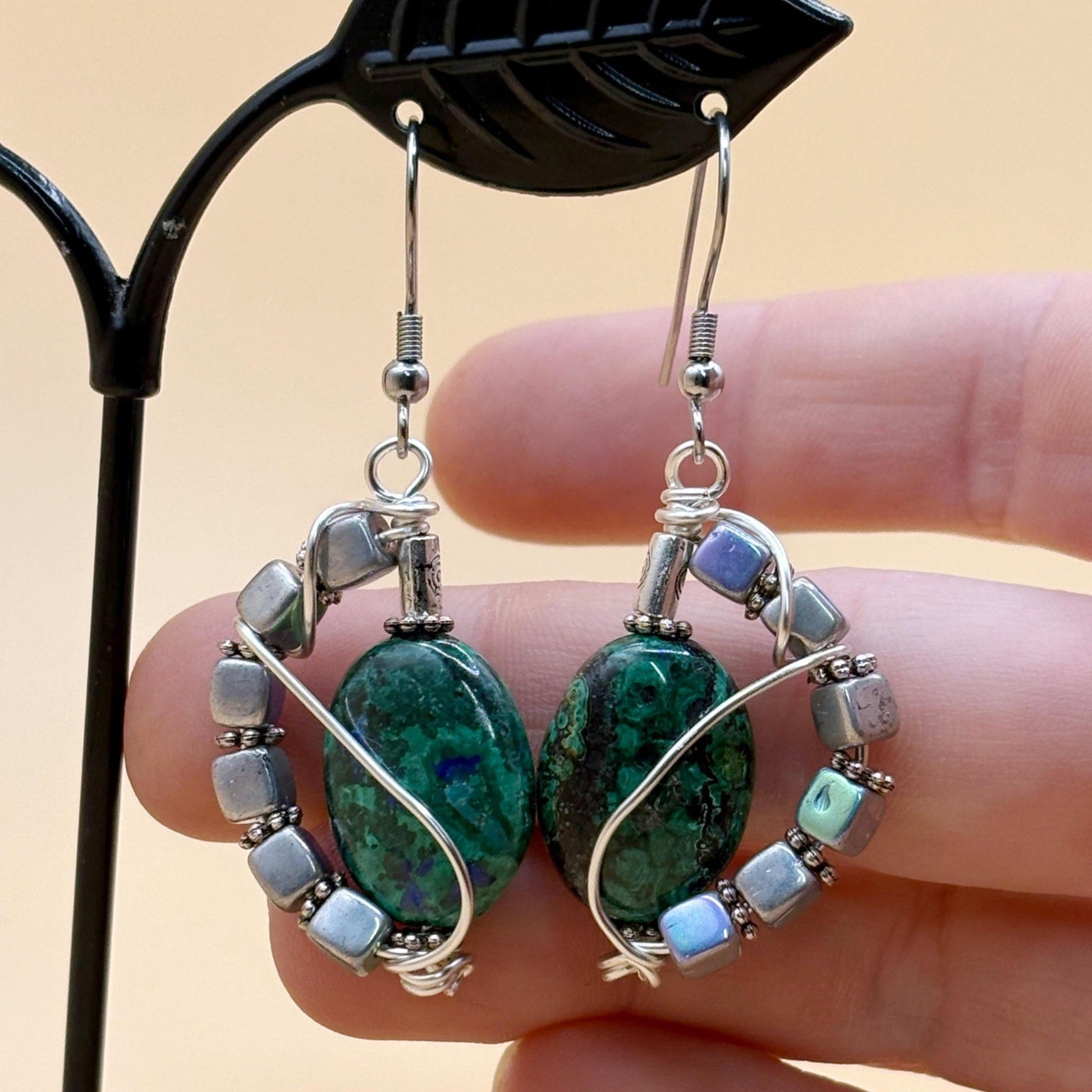 Pair of earrings with green stones and silver accents held by a hand against a beige background