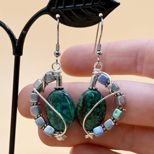 Pair of earrings with green stones and silver accents held by a hand against a beige background