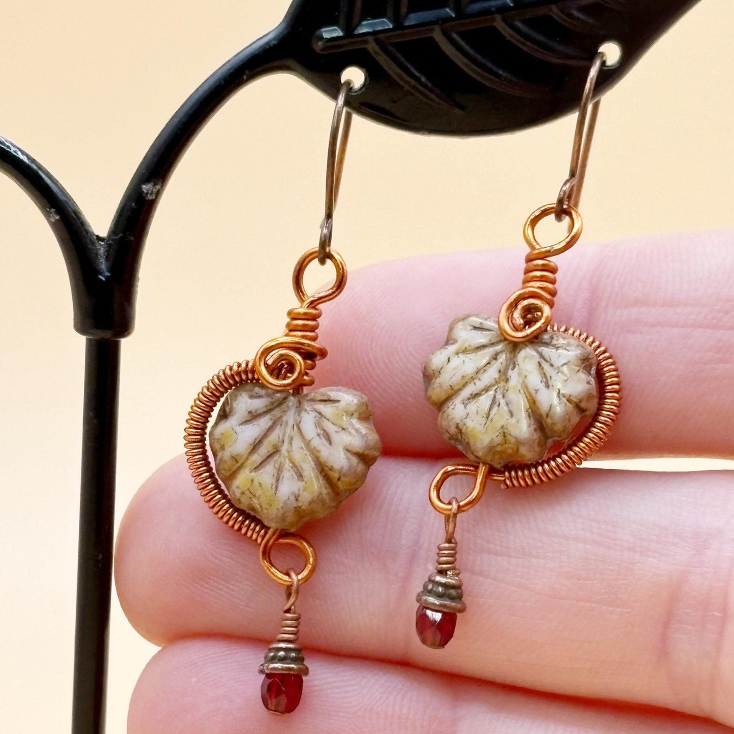 Pair of earrings with natural leaf shape glass beads and copper wirework held between fingers against a neutral background