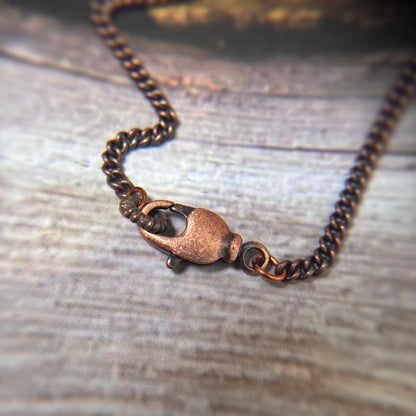 A close-up of a dark copper chain necklace with a matching lobster clasp, resting on a rustic, weathered gray wooden surface.