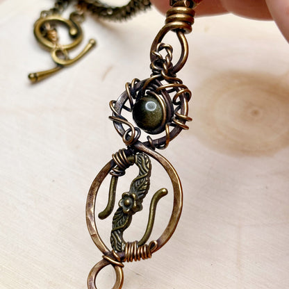 Close-up of a detailed bronze pendant with a central gold gemstone on a light background.