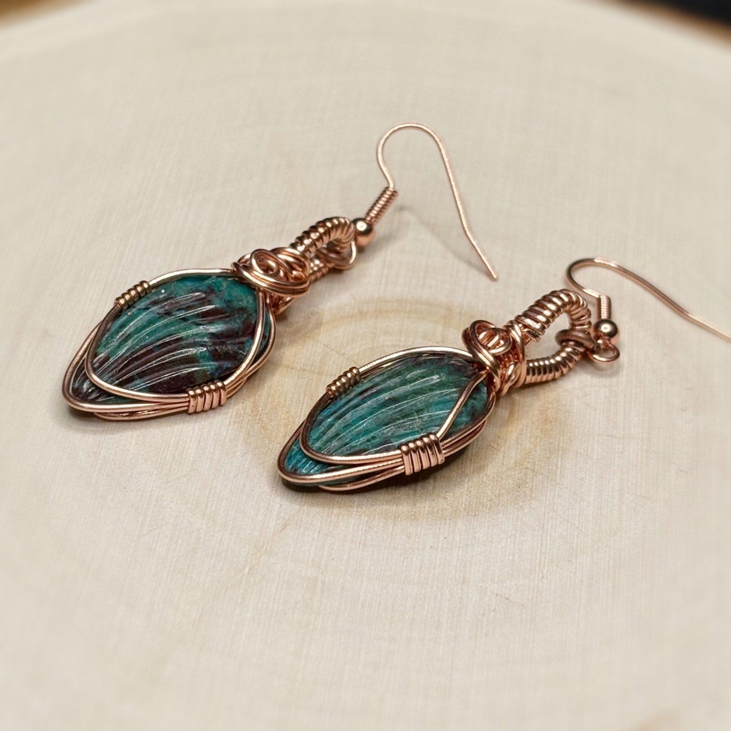 Pair of earrings with green stone and copper wire wrapping on a beige wood background