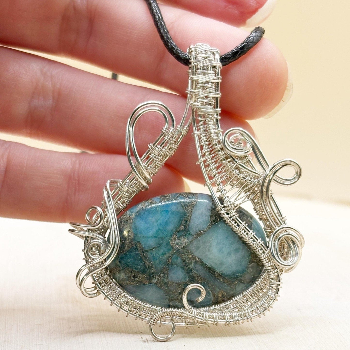 Silver wire-wrapped pendant with a blue stone held in a hand against a beige background