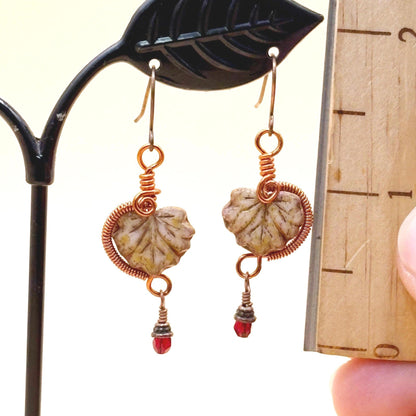 Pair of earrings with glass leaves and red bead details on a stand.