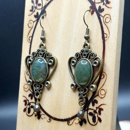 Pair of ornate earrings with green stones on a wooden stand with floral patterns.