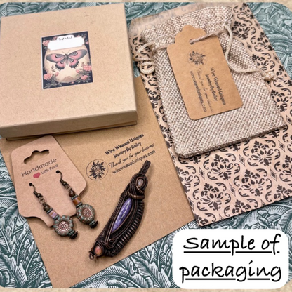A styled flat-lay showing craft packaging and accessories on a decorative patterned surface. Visible items include a kraft gift box with a butterfly label, a patterned fabric pouch with a kraft hangtag, a pair of dangling earrings on a card labeled handmade, and a dark beaded necklace on a kraft card.