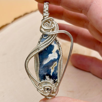 Silver wire-wrapped pendant with a blue and white stone held by a hand against a beige background