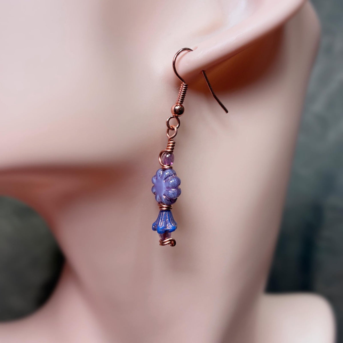 Copper wire wrap earrings with purple floral shaped beads hanging on an ear lobe