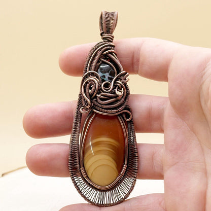 Hand holding a decorative pendant with a brown stone and intricate copper wirework.