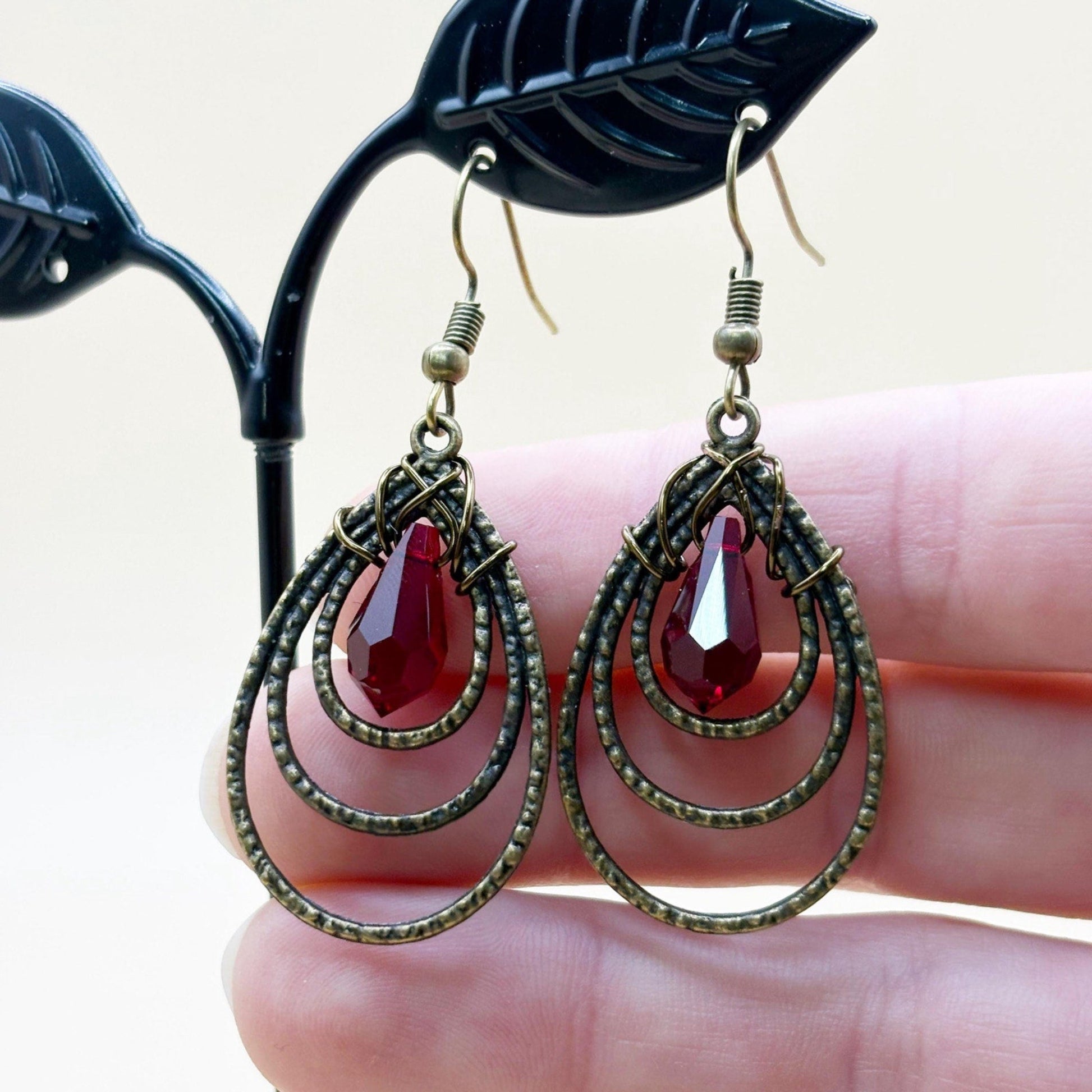 Pair of earrings with red teardrop-shaped stones held by a hand against a neutral background
