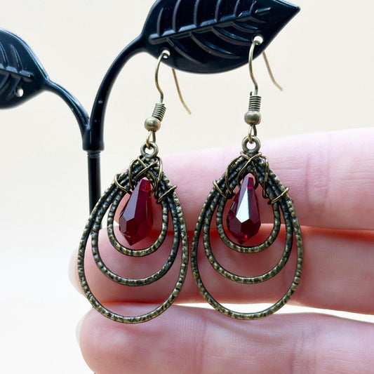 Pair of earrings with red teardrop-shaped stones held by a hand against a neutral background