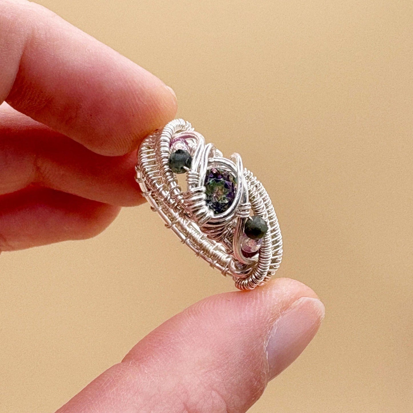 Silver ring with gemstones held between fingers against a beige background