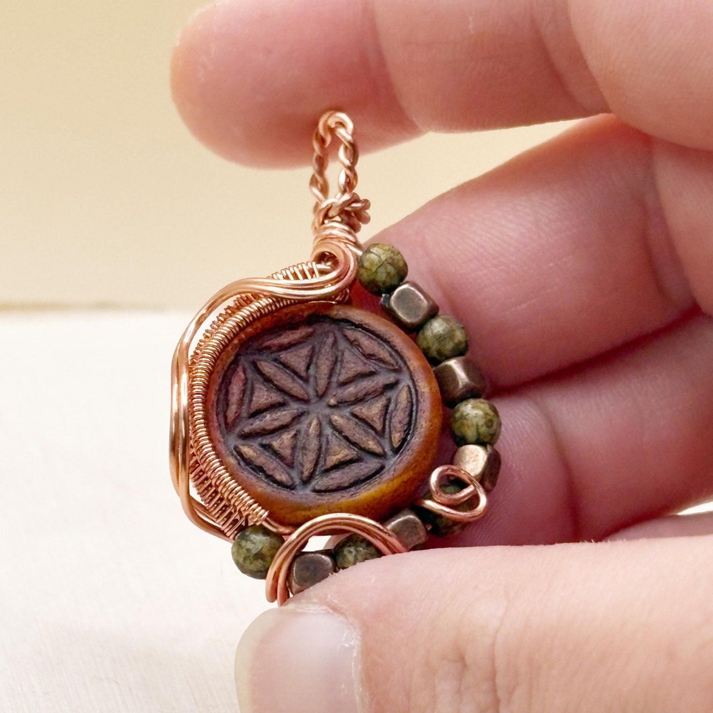 Ornate design wire wrap pendant jewelry copper with red focal flower of life and green beads being held in a hand.