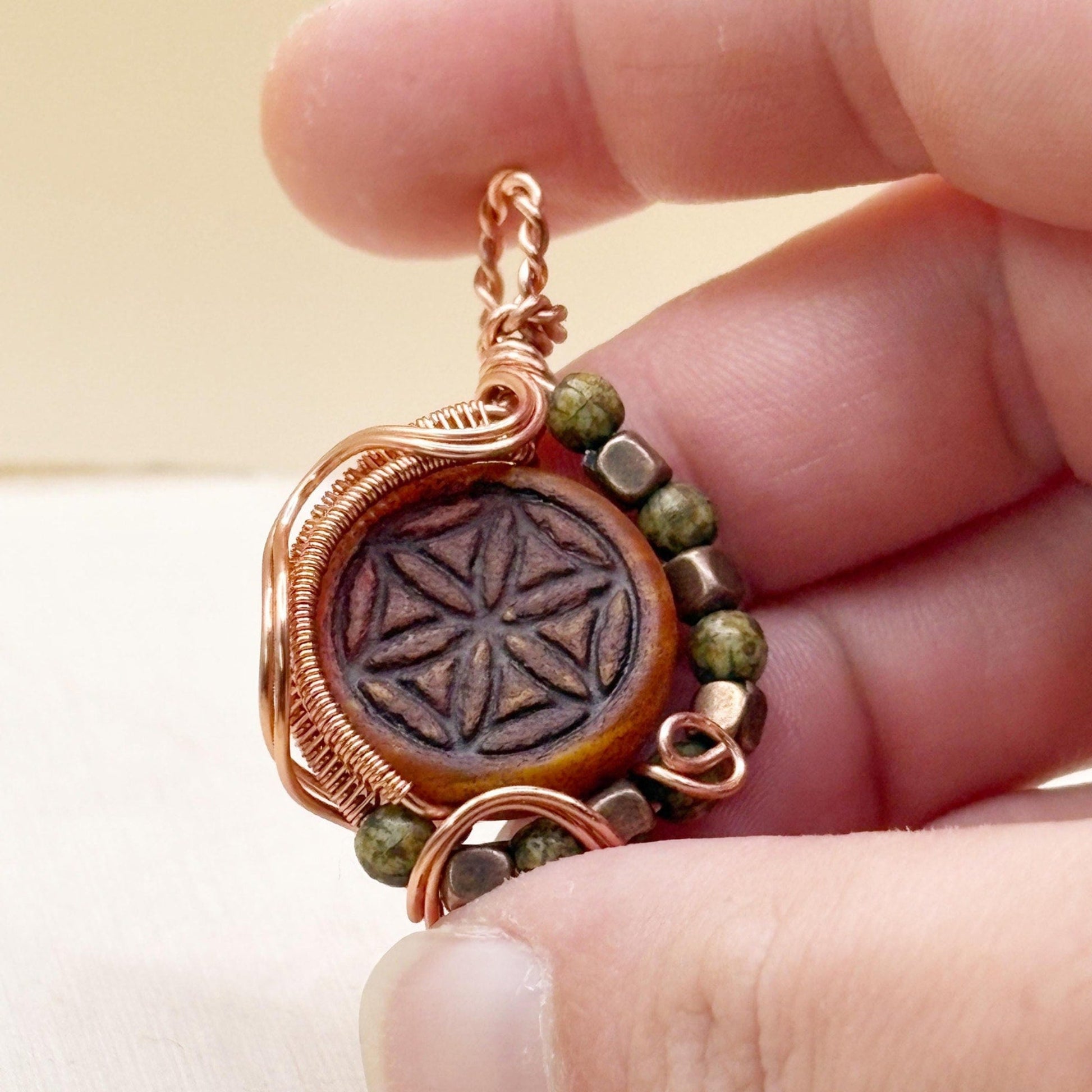 Ornate design wire wrap pendant jewelry copper with red focal flower of life and green beads being held in a hand.