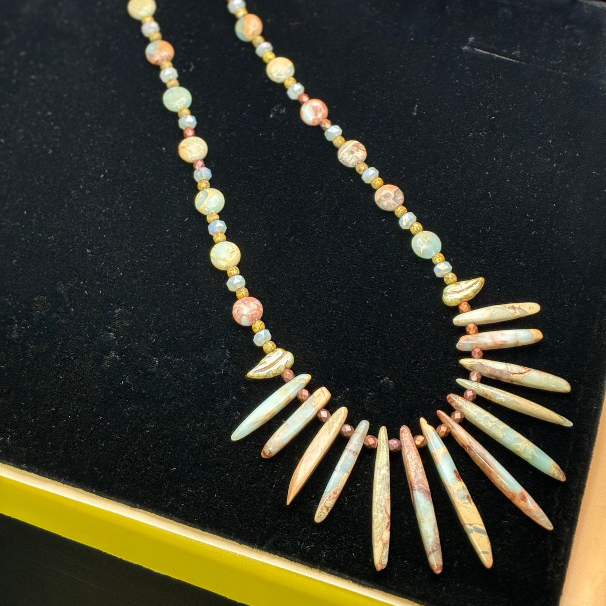 A beaded necklace displayed on a black velvet jewelry box. The necklace features a long strand of small marbled beads in pastel yellow, green, pink, and blue, with a large fan of elongated, tooth-like beads in cream, pink, and light blue-green at the bottom.