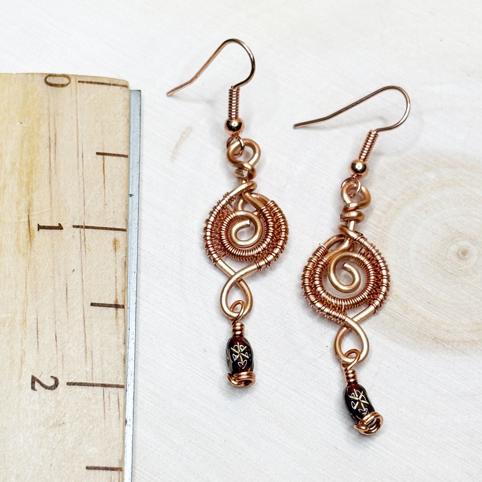 Pair of copper wire-wrapped earrings on a white background with a ruler for scale.