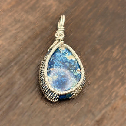 Teardrop-shaped pendant with blue stone and silver wire wrapping on a wooden surface
