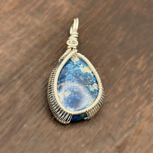 Teardrop-shaped pendant with blue stone and silver wire wrapping on a wooden surface