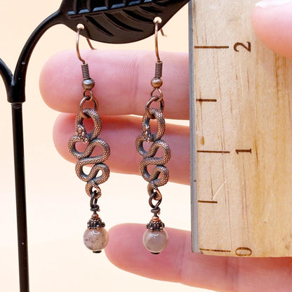 Pair of snake-style earrings held against a wooden ruler for size reference.