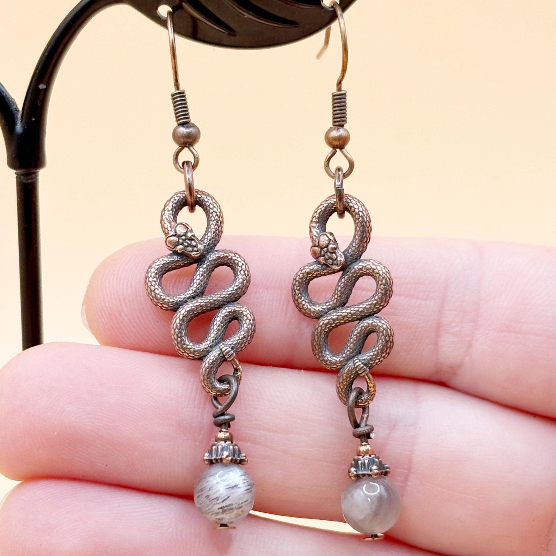 Pair of silver snake-shaped earrings with pearl-like beads held between fingers against a beige background