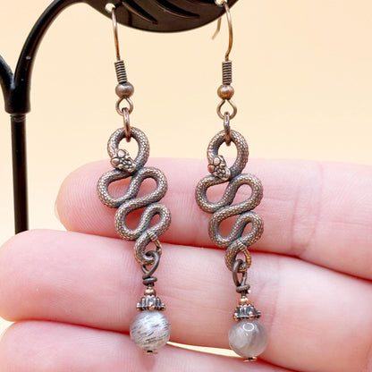Pair of silver snake-shaped earrings with pearl-like beads held between fingers against a beige background