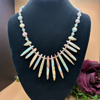 A statement necklace made of light blue and beige stone spikes with small pastel beads, displayed on a black velvet jewelry bust.