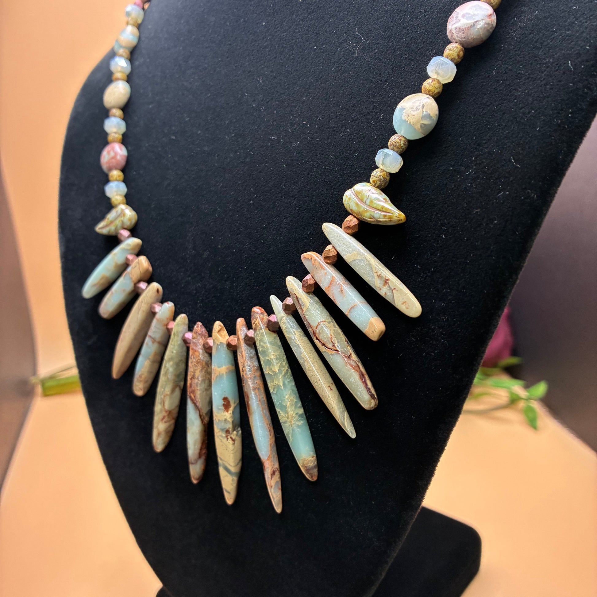 A beaded necklace displayed on a black jewelry bust. The necklace features a central cluster of elongated turquoise-blue and beige stone spacers, flanked by smaller round beads in blue, pink, and beige.