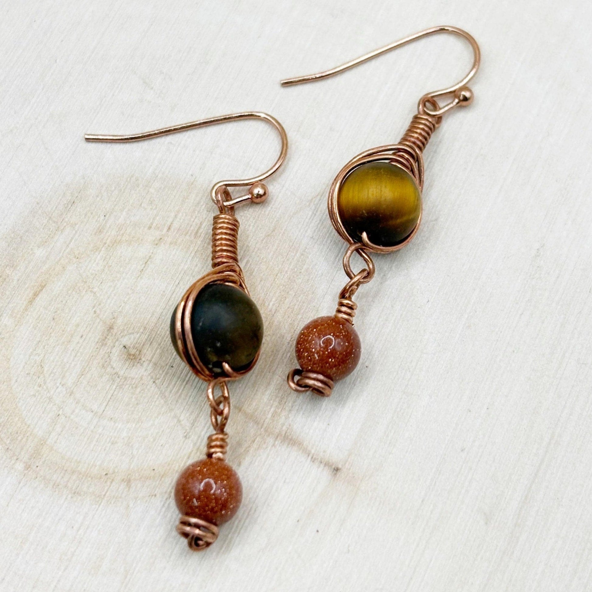 Pair of copper wire-wrapped earrings with brown and gold beads on a light wooden surface.