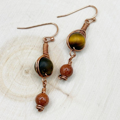 Pair of copper wire-wrapped earrings with brown and gold beads on a light wooden surface.