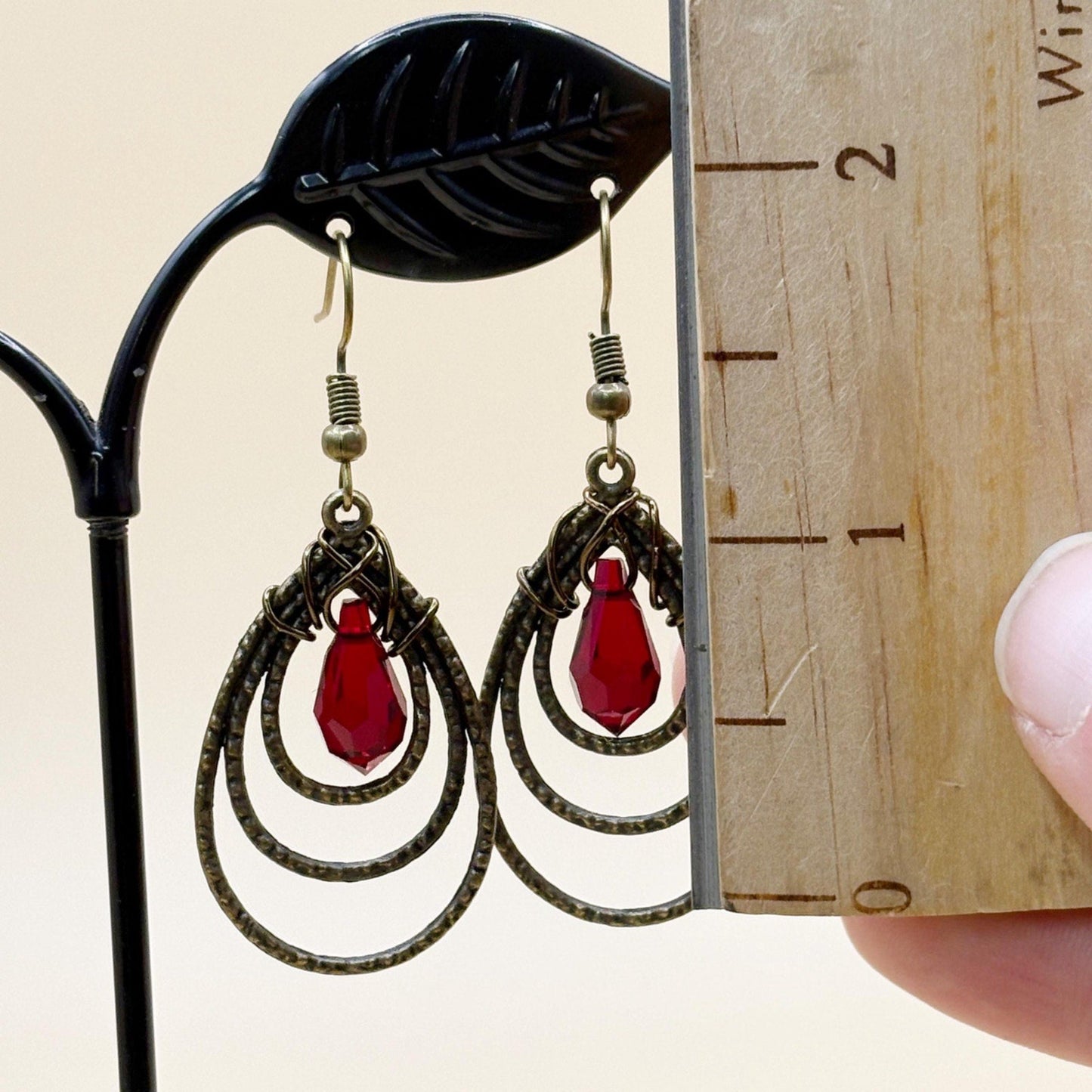 Pair of earrings with red teardrop-shaped stones on a stand next to a ruler for size reference.