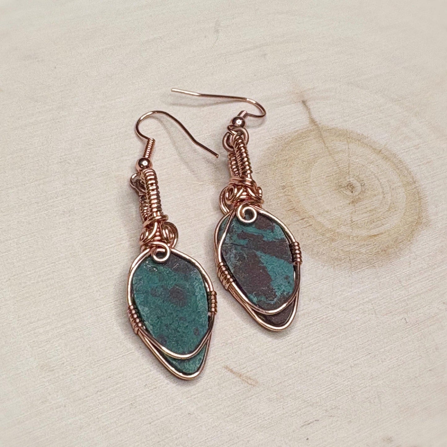 Pair of earrings with green stone and copper wire wrapping on a textured beige background