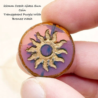 22mm Czech Glass Sun Coin with transparent purple color and bronze wash held between fingers.