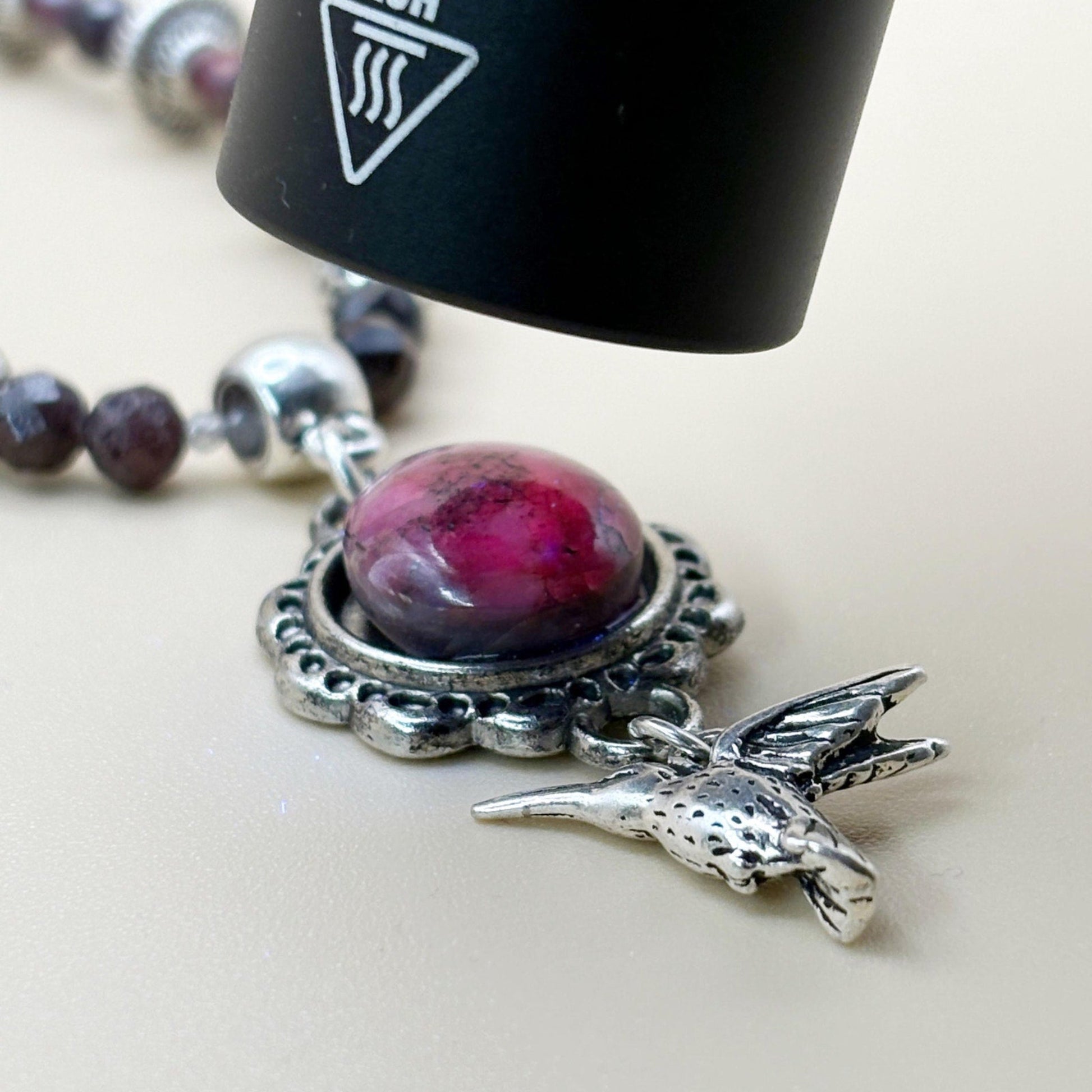 Close-up of a necklace with a pink bead and silver hummingbird charm on a light background