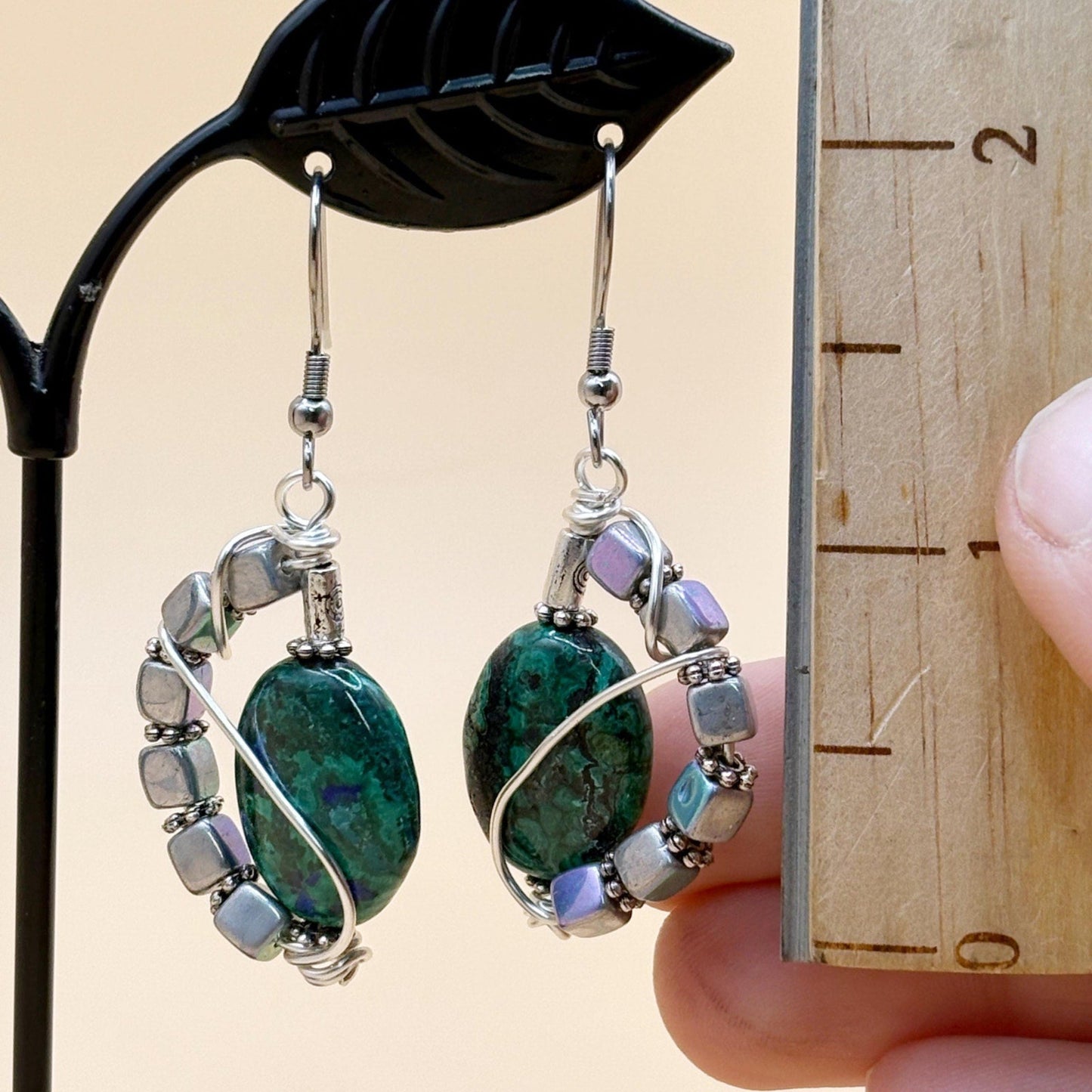 Pair of earrings with green and silver beads held next to a wooden ruler for size reference.