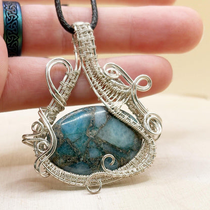Silver wire-wrapped pendant with a blue stone held between fingers on a light background
